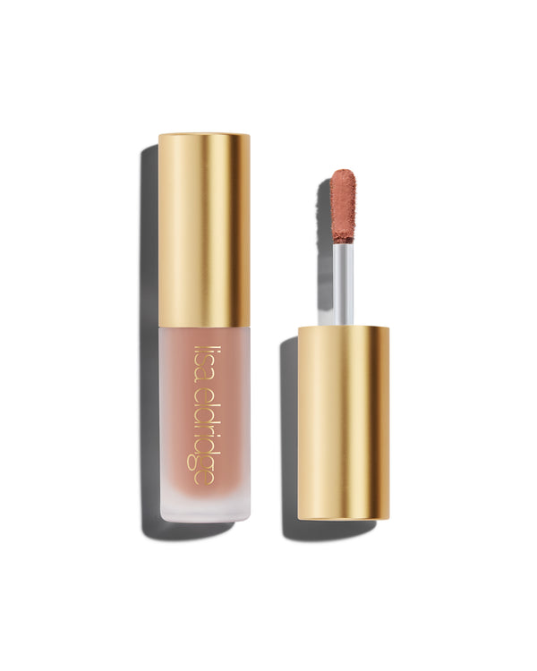 A closed bottle of Lisa Eldridge Liquid Silk Liquid Eyeshadow in peachy beige shade Irene, with a gold cap containing an applicator wand with product on it, against a white background.